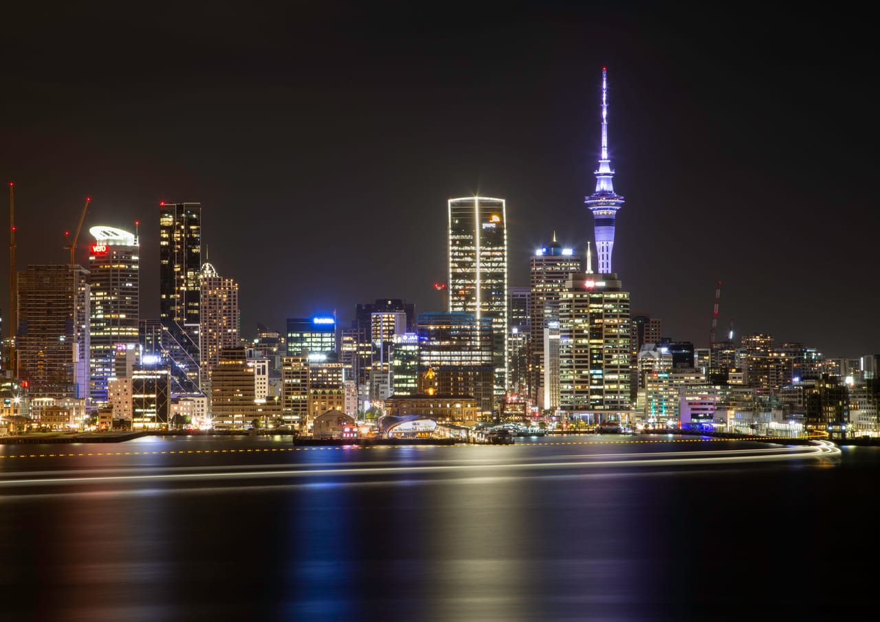 A wide view of Auckland's skyline illuminated at night with the Sky Tower visible.